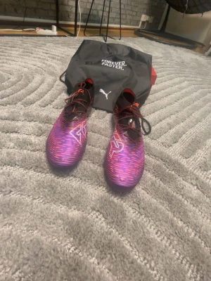 Puma lila fotbollsskor med väska. Kom med bud - Säljer ett par snygga fotbollsskor från Puma i lila och svart med röda detaljer och vita loggor. Skorna har snörning och en modern, strömlinjeformad design. Medföljer gör en svart väska med texten 'FOREVER. FASTER.' och Puma-logga. Perfekta för dig som vill sticka ut på planen.