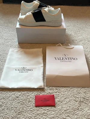 Valentino Garavani vita sneakers - Stilrena vita sneakers från Valentino Garavani med en bred svart remsa på sidan. Skorna har klassisk låg profil, vit sula med mönstrad undersida och är tillverkade i läder. Perfekta för dig som gillar exklusiv och modern design.