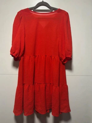 Röd volangklänning med puffärm - Trendig röd kortklänning med volanger och puffärmar. Klänningen har en lös passform och är tillverkad i ett strukturerat tyg som ger en cool look. Perfekt för dig som vill sticka ut med färg och volym.