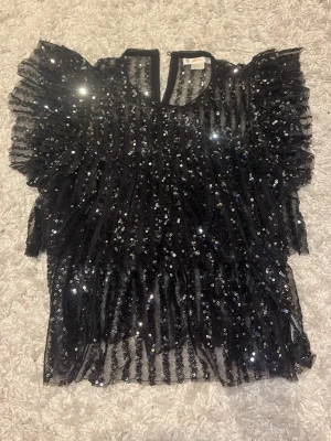 Svart glittrig topp med volangärmar - Supersnygg svart fest topp med glittriga paljetter och volangärmar från Gina tricot. Använd 1 gång, super bra skick. strl 140 = XXS 