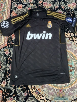 Real Madrid svart matchtröja Adidas - Snygg svart Real Madrid matchtröja från Adidas med guldiga detaljer och klubbmärke på bröstet. Tryckta sponsorloggor, Champions League-märke och UEFA Respect-märke på ärmarna. Tillverkad i lätt funktionsmaterial (Climacool) som andas.