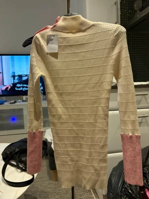  Tröja storlek xs - Ribbstickad tröja i beige med kontrasterande rosa muddar och axelparti. Hög hals och dekorativa guldknappar längs ena axeln ger en unik look. Långärmad och figurnära modell, perfekt för dig som gillar stilrena plagg med en twist.