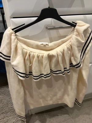 Off shoulder stickad tröja H&M - Krämvit stickad off shoulder-tröja från H&M med volang runt axlarna och svarta ränder som detalj. Långärmad modell i mjukt material, perfekt för en trendig och avslappnad look. Ribbstickade muddar vid ärmslut och nederkant.