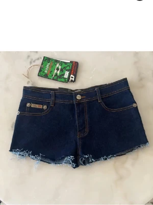 Mörkblå jeansshorts från Diesel - Säljer ett par mörkblå jeansshorts från Diesel med råa, fransiga benslut och kontrastsömmar. Shortsen har klassiska fem fickor och cool broderad detalj på bakfickorna. Perfekta för sommaren och riktigt snygga till sneakers eller sandaler.