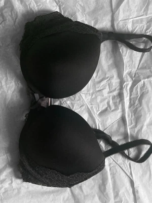 Svart spets-bh med vadderade kupor - Säljer en svart bh med vadderade kupor och dekorativ spets längs kanterna. Bh:n har justerbara axelband och klassisk knäppning bak. Materialet är en kombination av mjuk textil och spets som ger en elegant look.