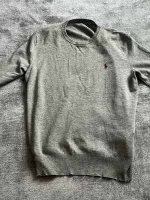 Grå stickad tröja från Ralph Lauren - Klassisk grå stickad tröja från Ralph Lauren med rund halsringning och diskret broderad logga i vinrött på bröstet. Tröjan har ribbade muddar vid ärmslut och nederkant. Perfekt till lager på lager och enkel att matcha.