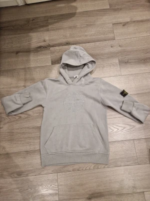 Grå hoodie från Stone Island - Snygg grå hoodie från Stone Island med broderad logga på bröstet och klassisk patch på ärmen. Hoodien har stor känguruficka framtill, ribbade muddar och en mjuk insida. Perfekt för en avslappnad streetwear-look.