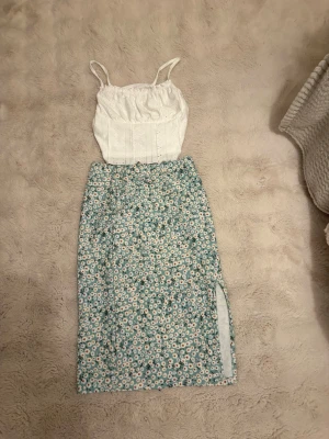 Vit blommig linnetopp med smala axelband och kjol med blommor  - Söt vit linnetopp med smala axelband och broderade detaljer. Toppen har en feminin och somrig känsla, perfekt till varma dagar. Den är ärmlös och har en lätt croppad passform som ger en trendig look. Snygg kjol till med fina blommor