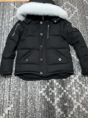 Svart pufferjacka med vit päls och svart  - Svart pufferjacka från Moose Knuckles med vit päls på huvan, silverfärgade dragkedjor och knappar. Jackan har en quiltad design, huva och logotyp på ärmen. Perfekt för kalla vinterdagar.