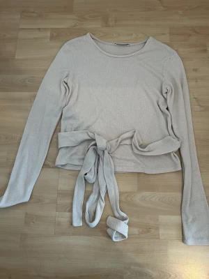 Beige croppad topp med knyt - Snygg beige croppad topp med långa ärmar och knyt i midjan. Toppen har en rund halsringning och är tillverkad i ett mjukt, stretchigt material som sitter bekvämt mot kroppen. Perfekt att styla med jeans eller kjol för en trendig look.