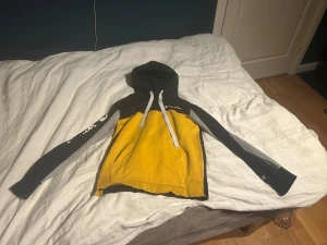 Gul och svart hoodie från Champion - Säljer en snygg hoodie från Champion i gul och svart med grå detaljer på ärmarna. Tröjan har huva med vita snören, magficka och långärmade armar. Perfekt för dig som gillar streetwear och vill sticka ut med färg.