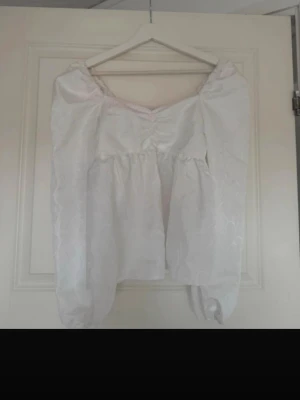 Vit topp med puffärmar - Säljer en vit topp med puffärmar. Toppen har en v-ringning framtill och är gjord i ett tyg med diskret blommönster. Perfekt för dig som gillar romantisk och trendig stil.