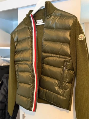 Olivgrön dunväst från Moncler -  JA ALLA SCANS OCH QR OCH TAGGAR FUNGERAR HELT RÄTTSnygg olivgrön dunväst från Moncler med stickade ärmar och hög krage. Västen har dragkedja framtill med Monclers klassiska röd-vit-blå rand och två fickor med dragkedja. Moncler-logga på ärmen. Perfekt för lager-på-lager och streetstyle.