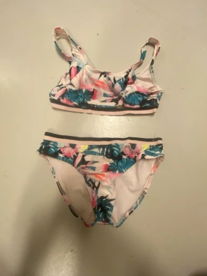Blommig bikini i pastellfärger - Säljer en snygg bikini med blommigt mönster i rosa, blått och vitt. Överdelen har breda axelband och sportig form, medan underdelen har en klassisk bikini-trosa med svart rand längs midjan. Perfekt för lata dagar på stranden eller poolen!