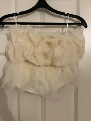 Vit bandeau topp i tyll från Market - Supersöt bandeau topp i vit tyll från Market. Toppen har en fluffig, voluminös design med lager av transparent tyg som ger en riktigt statement look. Perfekt för dig som vill sticka ut och älskar trendiga detaljer. Modellen är axelbandslös och croppad.