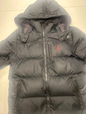 Svart dunjacka från Polo Ralph Lauren - Snygg svart dunjacka från Polo Ralph Lauren med huva och dragkedja framtill. Jackan har en röd logga broderad på bröstet, flera praktiska fickor med dragkedja och en puffig, varm design perfekt för kalla dagar. Klassisk och stilren look.Jackan är köpt på boozt för runt 3000kr. Inga defekter. Vid minsta frågor är det bara att skriva. Priser är ej fast.