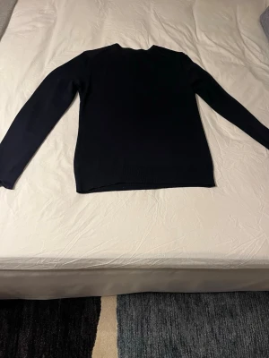 Svart stickad tröja J.Lindeberg - Snygg svart stickad tröja från J.Lindeberg med rund halsringning och diskret logga på bröstet. Tröjan har långa ärmar och ribbade muddar vid ärmslut och nederkant, 100% ull. 