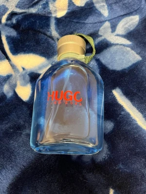 Hugo Boss blå parfymflaska - Säljer en blå parfym från Hugo Boss med en robust, rundad glasflaska och beige skruvkork med grön tygrem. Flaskan har röd logotyp på framsidan och sprayfunktion. Perfekt för dig som gillar stilrena och moderna dofter.