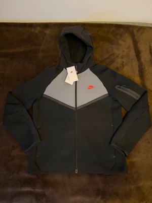 Nike Tech Fleece hoodie svart/grå - Nike Tech Fleece hoodie i svart och grått med dragkedja framtill och huva. Har en röd Nike-logga på bröstet och en praktisk ficka med dragkedja på ärmen. Tillverkad i mjukt och lätt material som ger en modern och sportig look. Helt ny aldrig använd med prislapp. Ny pris på cirka 1500kr