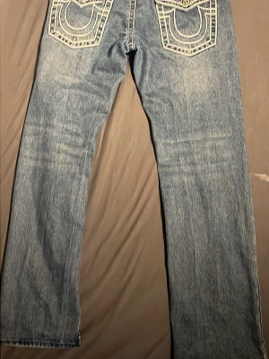 Blå True Religion jeans med kontrastsömmar - Säljer ett par blå jeans från True Religion med raka ben och tydliga vita kontrastsömmar. Baksidan har de ikoniska stora fickorna med broderade detaljer. Klassisk denimlook i 100% bomull, perfekt för dig som gillar streetwear och vill sticka ut.