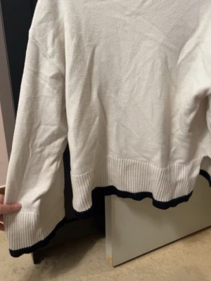 Vit stickad tröja med svarta detaljer - Vit stickad tröja med långa ärmar och ribbstickade muddar. Tröjan har svarta vågiga detaljer längs nederkant och ärmslut för en cool kontrast. Perfekt för dig som gillar enkel men snygg stil.