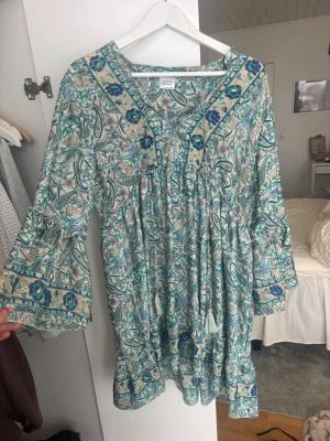 Sommar klänning - Supersöt kortklänning från Moeva med blommigt mönster i blått, grönt och beige. Klänningen har v-ringning, volangdetaljer och vida trekvartsärmar. Tillverkad i mjuk bomull, perfekt för sommarens alla dagar. Köpt i Spanien 