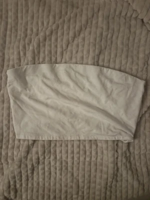 Vit bandeau topp L/XL - En enkel och stilren vit bandeau topp i mjukt och stretchigt material. Perfekt att bära under en skjorta eller som den är. Toppen har en elastisk kant upptill och nertill för att sitta bekvämt på plats. Passar dig som gillar minimalistisk stil.