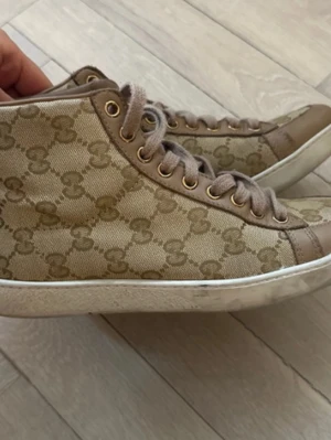 Beige höga sneakers från Gucci - Snygga höga sneakers från Gucci i beige canvas med det klassiska GG-mönstret och detaljer i ljusbrunt läder. Skorna har vita sulor och guldfärgade öljetter för snörningen. Perfekta för dig som gillar exklusiv streetstyle.