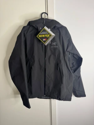 Svart GORE-TEX regnjacka Arc'teryx storlek M - Snygg svart regnjacka från Arc'teryx med GORE-TEX-material som håller dig torr i alla väder. Jackan har huva, hel dragkedja framtill och två praktiska sidofickor med dragkedja. Perfekt för dig som vill ha en stilren och funktionell jacka.