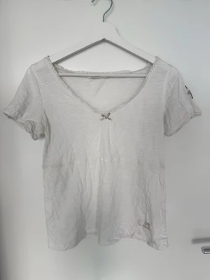 Topp från Odd Molly - Säljer en vit t-shirt från Odd Molly med v-ringning och liten rosett framtill. T-shirten har korta ärmar. Tillverkad i mjuk bomull och har fina spetsdetaljer vid halsen.