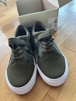 Klassiska kamouflage sneakers barn Nya i kartong - Snygga sneakers från Warp i militärgrönt med kamouflagemönster. Skorna har både kardborreband och elastiska skosnören för enkel påtagning. Ovandelen är i textil och mockaimitation, sulan är vit och i gummi. Perfekta för en cool och avslappnad stil.