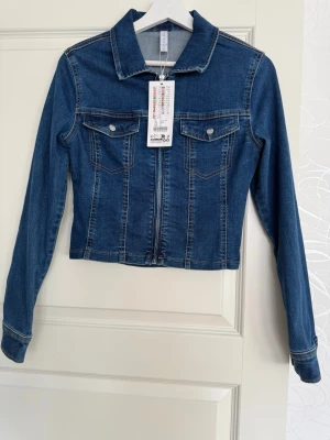 Mörkblå jeansjacka med dragkedja - Snygg mörkblå jeansjacka med dragkedja framtill och två bröstfickor med lock och knapp. Den är i storlek s men passar även xs då den är liten i storlek 