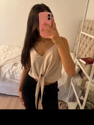 Beige omlottopp från Zara - Snygg beige omlottopp från Zara med knyt framtill och v-ringad hals. Toppen är ärmlös och har en mjuk, lätt glansig yta som ger en stilren look. Sitter som xs 