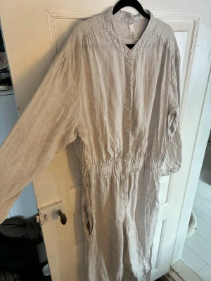 Beige jumpsuit i linne från kay/day - Säljer en beige jumpsuit från kay/day i storlek L. Den är tillverkad i linne, har långa ärmar, knäppning framtill och elastisk midja. Perfekt för dig som gillar en avslappnad och stilren look. Fickor på sidorna och enkel design.