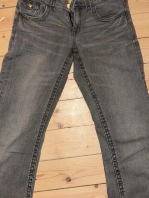 Grå raka jeans med slitningar - Säljer ett par grå jeans med raka ben och diskreta slitningar. Klassisk femficksmodell med knappgylf och detaljer i mässing. Jeansen har en avslappnad passform och är tillverkade i mjuk denim för skön känsla.