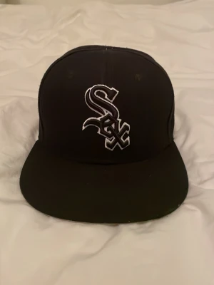 Svart New Era White Sox keps - Säljer en svart keps från New Era med Chicago White Sox-logga broderad i vitt framtill. Klassisk flat brim och broderad New Era-logga på sidan. Snygg streetwear vibe och passar perfekt till en avslappnad stil. Den enda skavanken med kepsen är att den saknar knoppen på toppen.