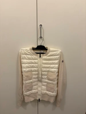 Vit cardiganjacka från Moncler - Snygg vit cardiganjacka från Moncler med quiltad, vadderad framsida och stickade ärmar. Rund halsringning, två fickor framtill och dragkedja. Ikonisk Moncler-logga på ärmen. Perfekt för lager-på-lager och en clean look.