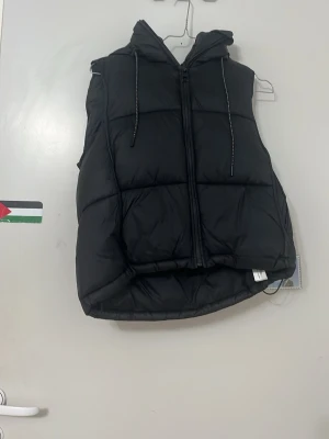 Svart dunväst med huva - Snygg svart dunväst med dragkedja och stor huva. Västen har quiltad design och dragsko i huvan för extra stilpoäng. Perfekt att slänga över en hoodie för en chill look. Materialet är vadderat och ger en puffig känsla.