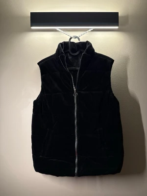 Svart dunväst med dragkedja - Stilren svart dunväst med hög krage och silverfärgad dragkedja framtill. Västen har två fickor på sidorna och quiltad design som ger en schysst look. Perfekt att slänga över en hoodie eller tröja för extra värme. Materialet är mocka och ger en lyxig känsla.