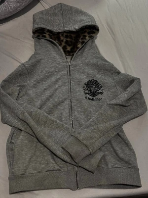 Grå zip-hoodie med leopardfoder - Snygg grå zip-hoodie från Challenge med leopardmönstrat foder i huvan. Dragkedja framtill, ribbade muddar och tryckt svart logga på bröstet. Perfekt för dig som gillar streetwear och vill ha något unikt i garderoben.