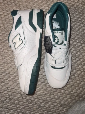 New Balance 550 vita/gröna sneakers - Snygga New Balance 550 sneakers i vitt och mörkgrönt läder med klassisk retrodesign. Skorna har perforerade detaljer, gröna loggor och matchande gröna insidor. Perfekta för dig som gillar stilrena och sportiga sneakers.