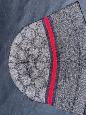 Grå mössa med röd och marinblå rand - Snygg grå mössa från Gucci med diskret GG-mönster över hela ytan. Mössan har en klassisk passform och pryds av en marinblå och röd rand runt om. Tillverkad i mjukt stickat material som håller dig varm och stilren.