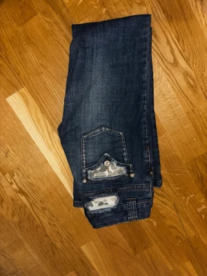 Mörkblå jeans med detaljer - Säljer ett par mörkblå jeans från Girls med snygga slitningar och coola detaljer som glittriga hällor och mönstrade fickor. Jeansen har dubbelknäppning fram och klassisk femficksdesign. Perfekta för dig som vill ha något extra i vardagen.