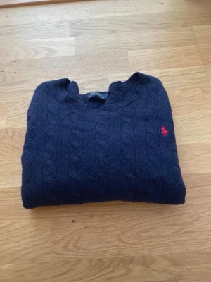 Mörkblå kabelstickad tröja Polo Ralph Lauren - Mörkblå kabelstickad tröja från Polo Ralph Lauren med klassisk röd logga broderad på bröstet. Tröjan har rund halsringning och ribbade muddar vid ärmslut och nederkant. Perfekt för lager på lager och riktigt snygg till jeans.