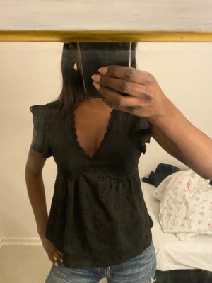 Svart blus med volangärmar och spets - Söt svart blus med v-ringning, volangdetaljer på ärmarna och spetskant längs urringningen. Blusen har en luftig passform och små broderade prickar i tyget som ger en extra touch. Perfekt att styla med jeans eller kjol.