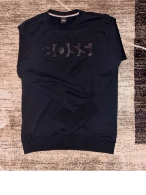 Svart sweatshirt från BOSS - Snygg svart sweatshirt från BOSS med stor logga i blankt material över bröstet. Tröjan har rund halsringning, långa ärmar och ribbade muddar vid ärmslut och nederkant. Perfekt för en stilren och avslappnad look.