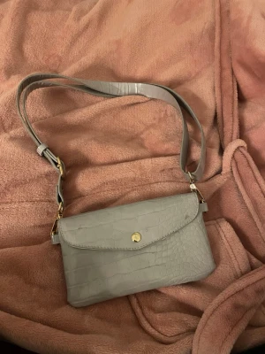 Grå axelväska i crocopräglat från Lindex - Snygg grå axelväska från Lindex med crocopräglat mönster och justerbar axelrem. Väskan har guldfärgade detaljer och stängs med tryckknapp. Insidan är ljus och rymlig, perfekt för det viktigaste. Enkel och stilren design som passar till många outfits.