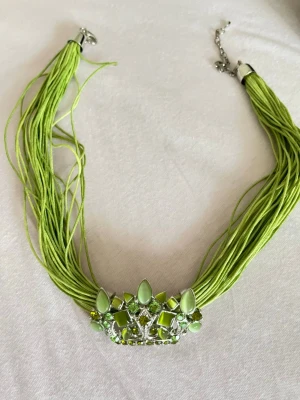 Grönt statementhalsband med pärlor - Unikt halsband med flera gröna trådar och ett stort dekorerat smycke framtill med gröna pärlor och stenar i olika former. Halsbandet har silverfärgade detaljer och justerbart lås. Perfekt för dig som vill sticka ut med färg och cool design.