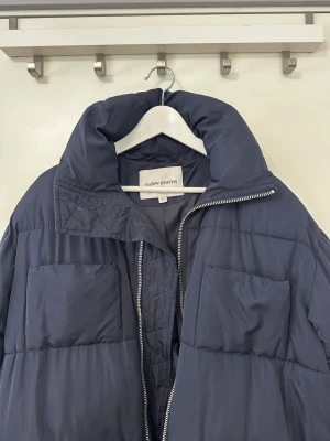 CARIN WESTER marinblå vinterjacka - Oversized, passar S med. En marinblå Carin Wester Hailey pufferjacka med en hög krage och dragkedja framtill. Jackan har två stora fickor framtill med lock och en avslappnad passform som ger en modern silhuett. Den är designad för att vara både funktionell och stilren, perfekt för kyligare väder.