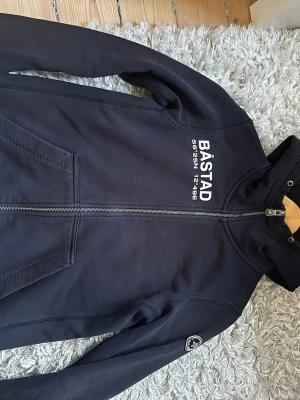 Pelle P Hoodie  - Snygg marinblå hoodie med dragkedja och vit text 'BÅSTAD' samt koordinater på bröstet. Huva och två fickor framtill. Liten patch på ärmen. Perfekt för dig som gillar stilren och sportig look. Mjuk bomullsblandning för skön känsla.